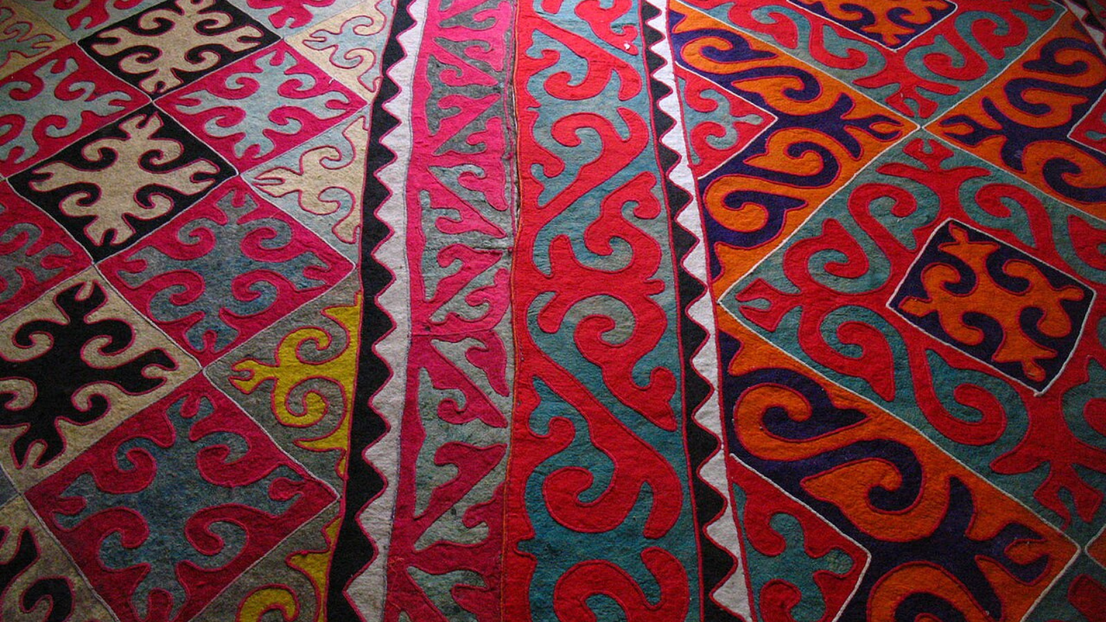 A colorful shyrdak, a type of rug, with geometric square and diamond patterns filled with spiraling and curvy abstract shapes in bright pink, red, navy, yellow, teal and light and dark blues.